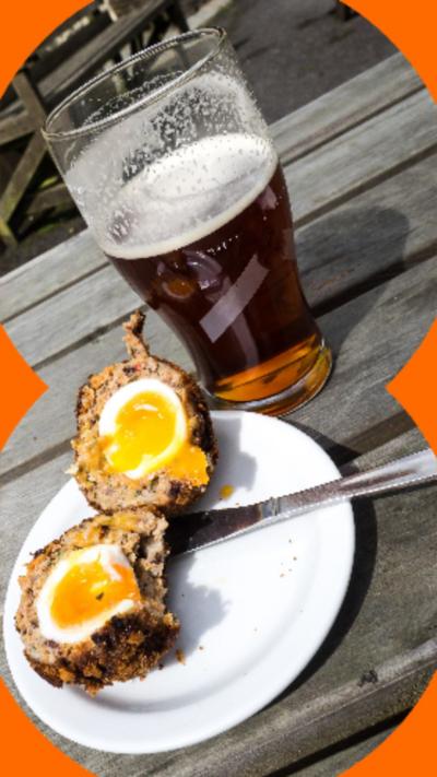 Image of a Scotch egg cut in half, next to a pint on an outdoor picnic table.