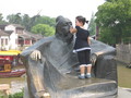 A small kid covering a wise-looking statue's mouth. It is cute.